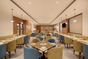 Uma sala de conferências de hotel com mesas, cadeiras, tela de projetor e área de cozinha.