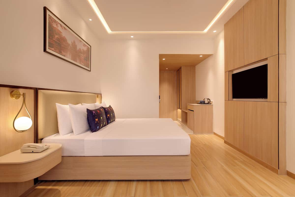 A modern hotel room with a bed, bedside table, lamp, TV, and a small kitchenette.