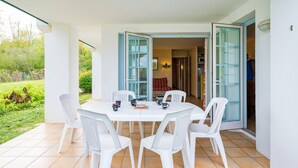 A patio with a round table and white chairs, open doors leading to an interior, and a garden view.