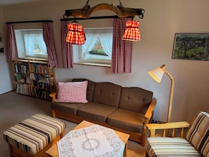 Una habitación acogedora con un sofá marrón, una estantería de madera, un reposapiés estampado y una mesa con mantel decorativo.