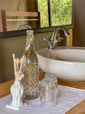 Une salle de bain avec un lavabo blanc, une bouteille en verre ornée d’un motif en zigzag et des verres transparents posés sur un tissu blanc.