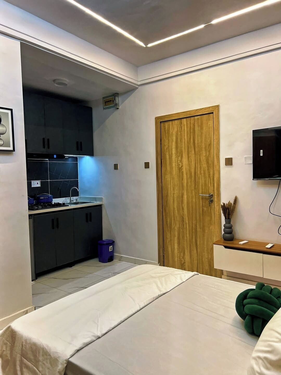 Um quarto com cama, porta de madeira, televisão e uma pequena cozinha.