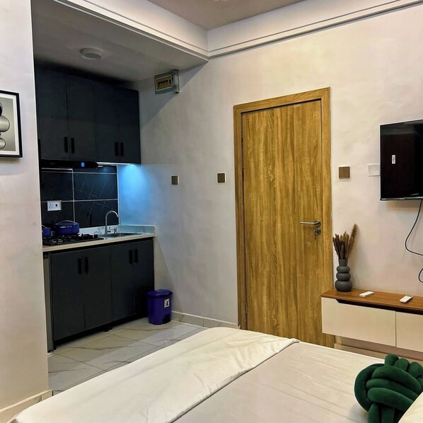 Une petite pièce comprenant un coin cuisine, un lit, une porte en bois et un téléviseur fixé au mur.