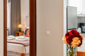 A hotel room with a bed, a bedside table with a lamp, a wall-mounted TV, and a kitchen area with a refrigerator and a coffee machine. - Empress 1A by Homie (Funchal)