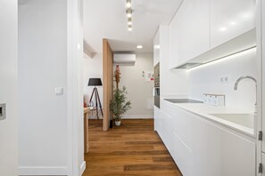 A modern kitchen with white cabinetry, a wooden floor, and a small dining area with a lamp and plant. - Empress 3F by Homie (Funchal)