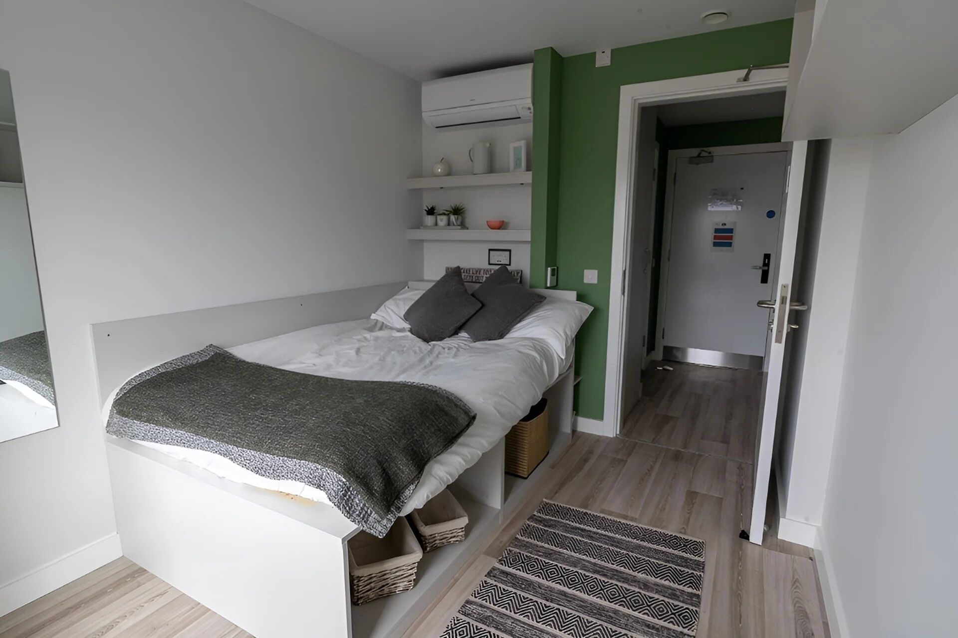 Un lit simple avec une couverture grise, une table de chevet, une petite étagère avec des objets personnels et un tapis à motifs sur un plancher en bois.