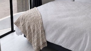 A bedroom with a large window, a bed with white bedding, and a beige throw blanket. - Zerilith Haven self catering (Swakopmund)