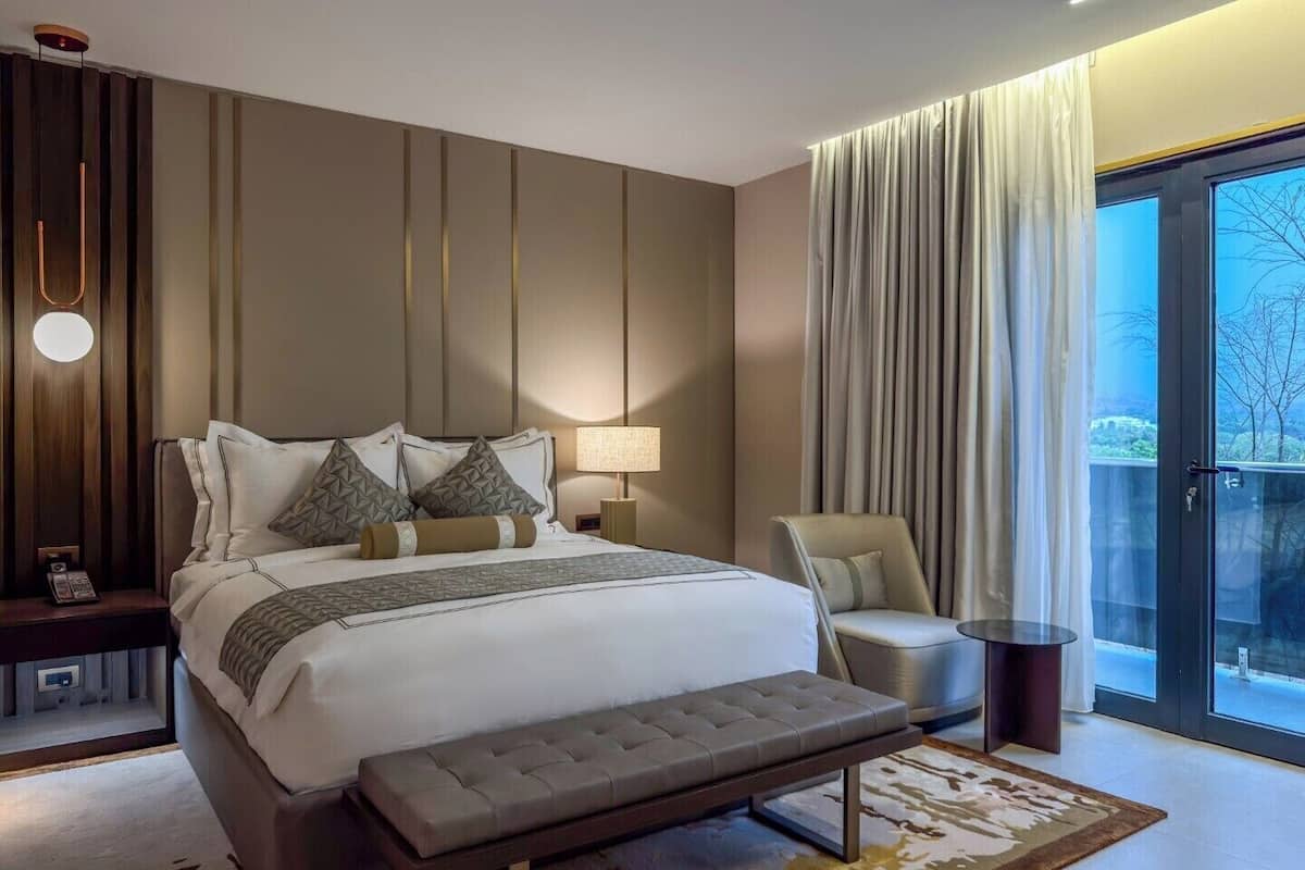 A modern hotel room with a large bed, a bench, a chair, a bedside table, a lamp, and a view of the outdoors.