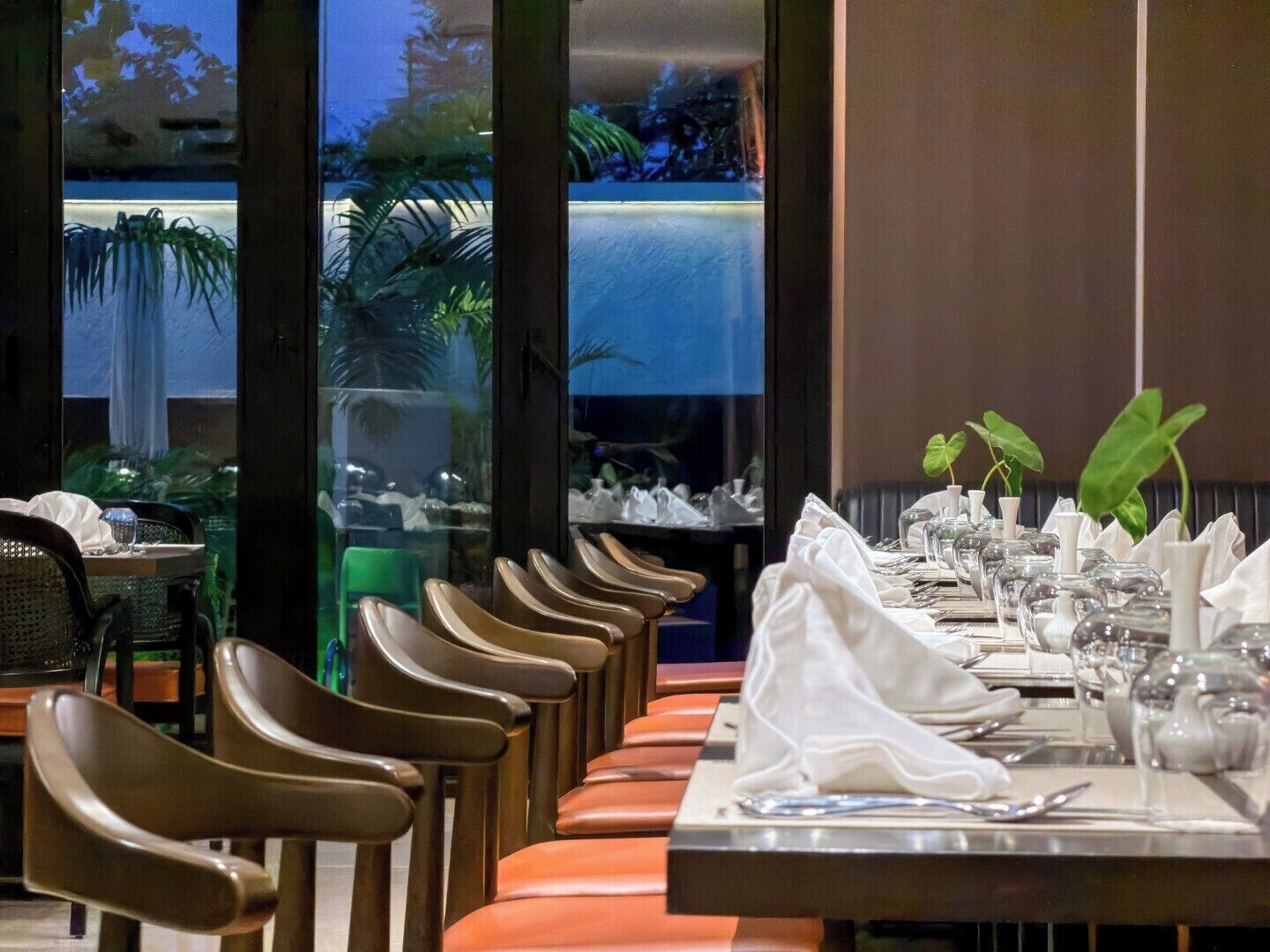A restaurant with a long dining table set for multiple guests, chairs aligned on both sides, and a view of a water feature outside.