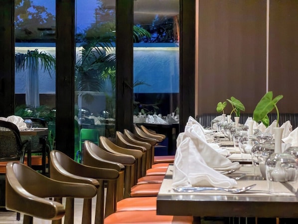 A restaurant with a long dining table set for multiple guests, chairs aligned on both sides, and a view of a water feature outside.