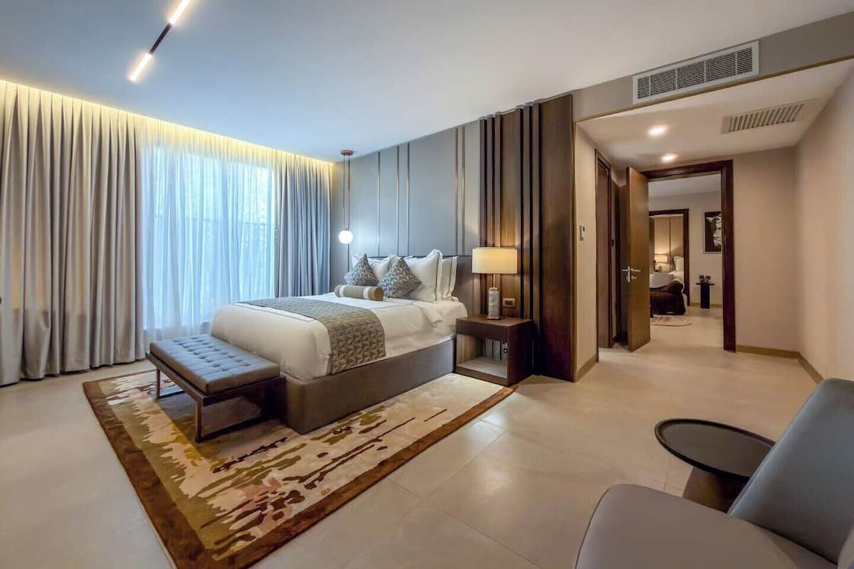 A modern hotel room with a large bed, a bedside table with a lamp, a chair, and a view of the hallway.