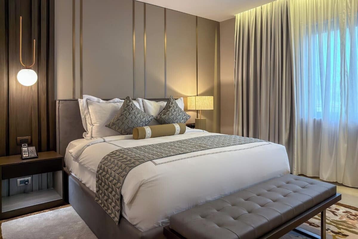 A modern hotel room with a large bed, bedside tables, a bench, and a view of the outdoors.