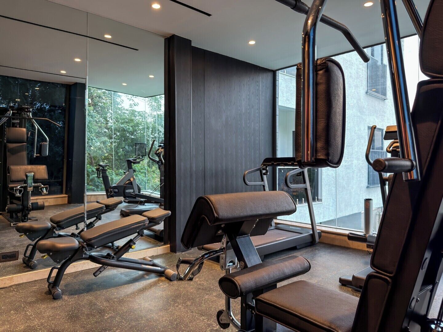 A modern gym with exercise equipment, large windows, and a view of greenery.