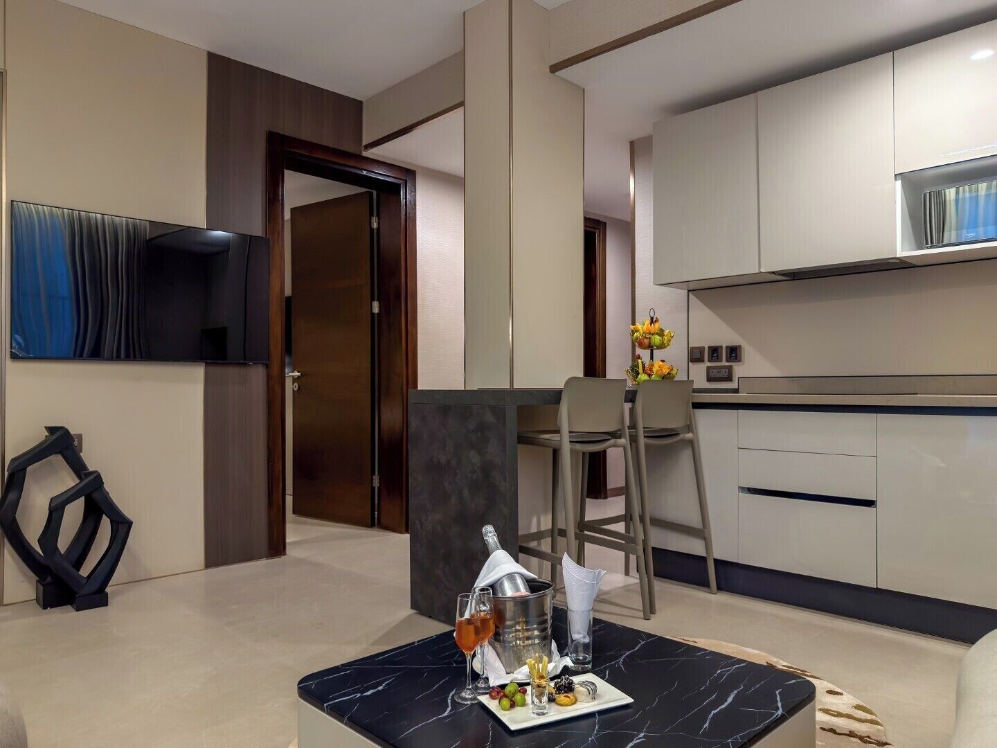 A modern hotel room with a dining area, a TV mounted on the wall, and a kitchen with cabinets.