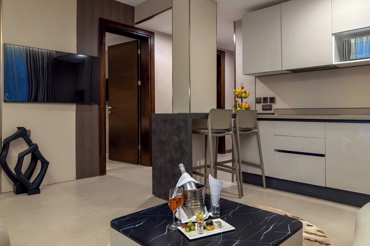 A modern hotel room with a dining area, a TV mounted on the wall, and a kitchen with cabinets.