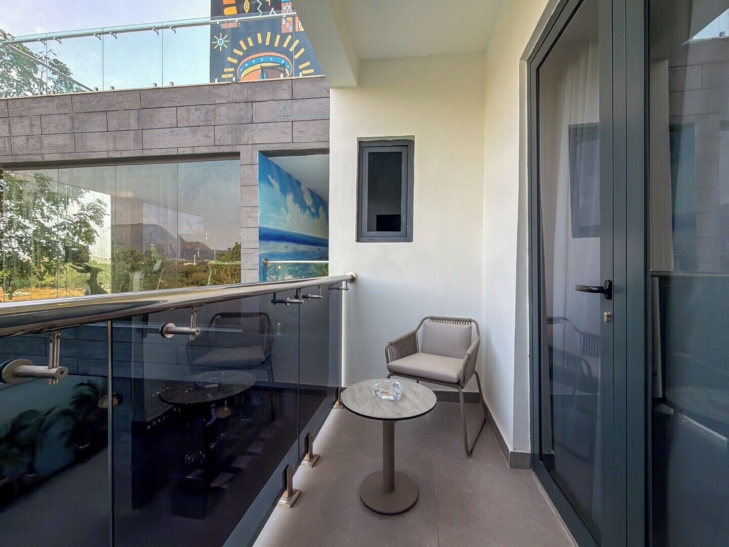 Ein Balkon mit Glastrennwand, einem kleinen runden Tisch, einem gepolsterten Stuhl und einer Tür auf der rechten Seite.