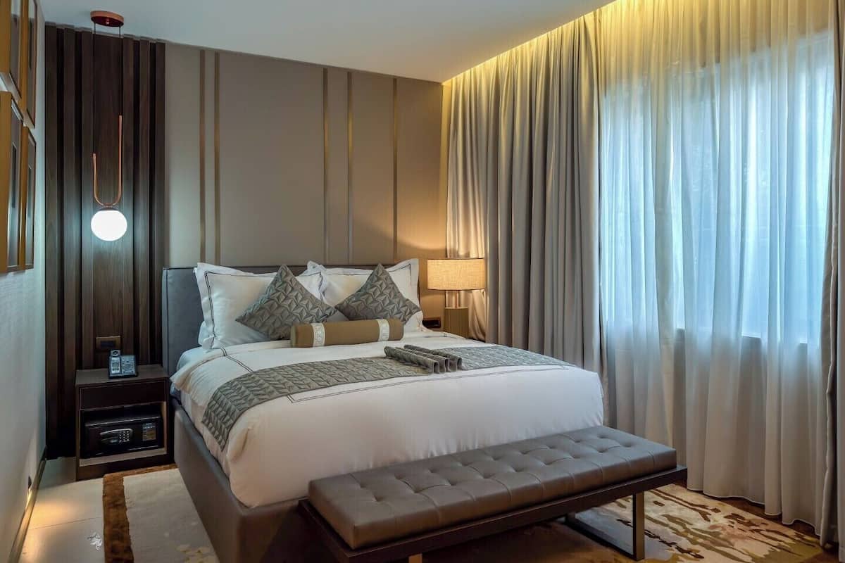 A hotel room with a large bed, a bedside table with a lamp, a bench, and a view of the outside through sheer curtains.
