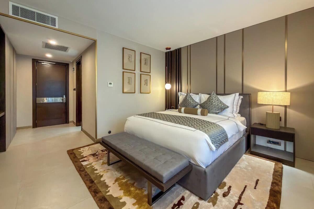 A modern hotel room with a large bed, a bench, a bedside table, and a wall-mounted lamp.