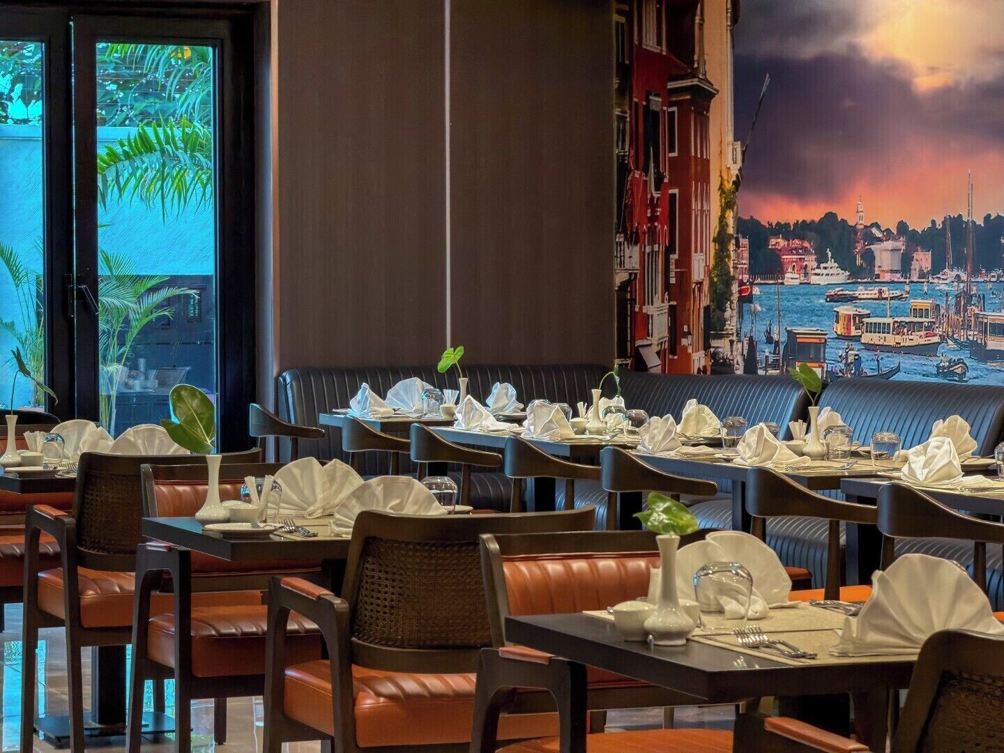 A restaurant with multiple tables set for dining, a large window with a view of the city, and a mural depicting a cityscape.