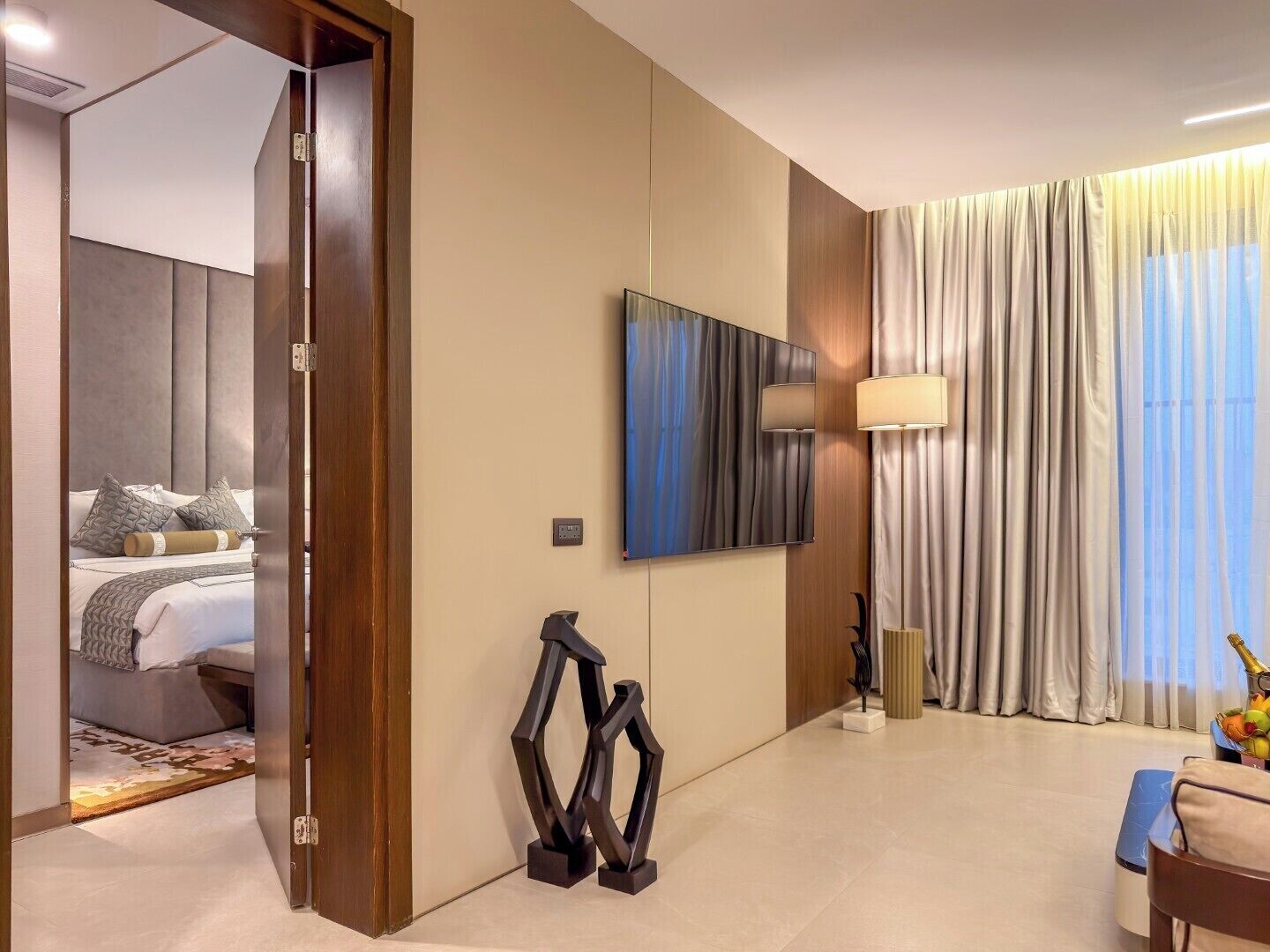 A hotel room with a bed, a flat-screen TV, a lamp, and a large window with curtains.