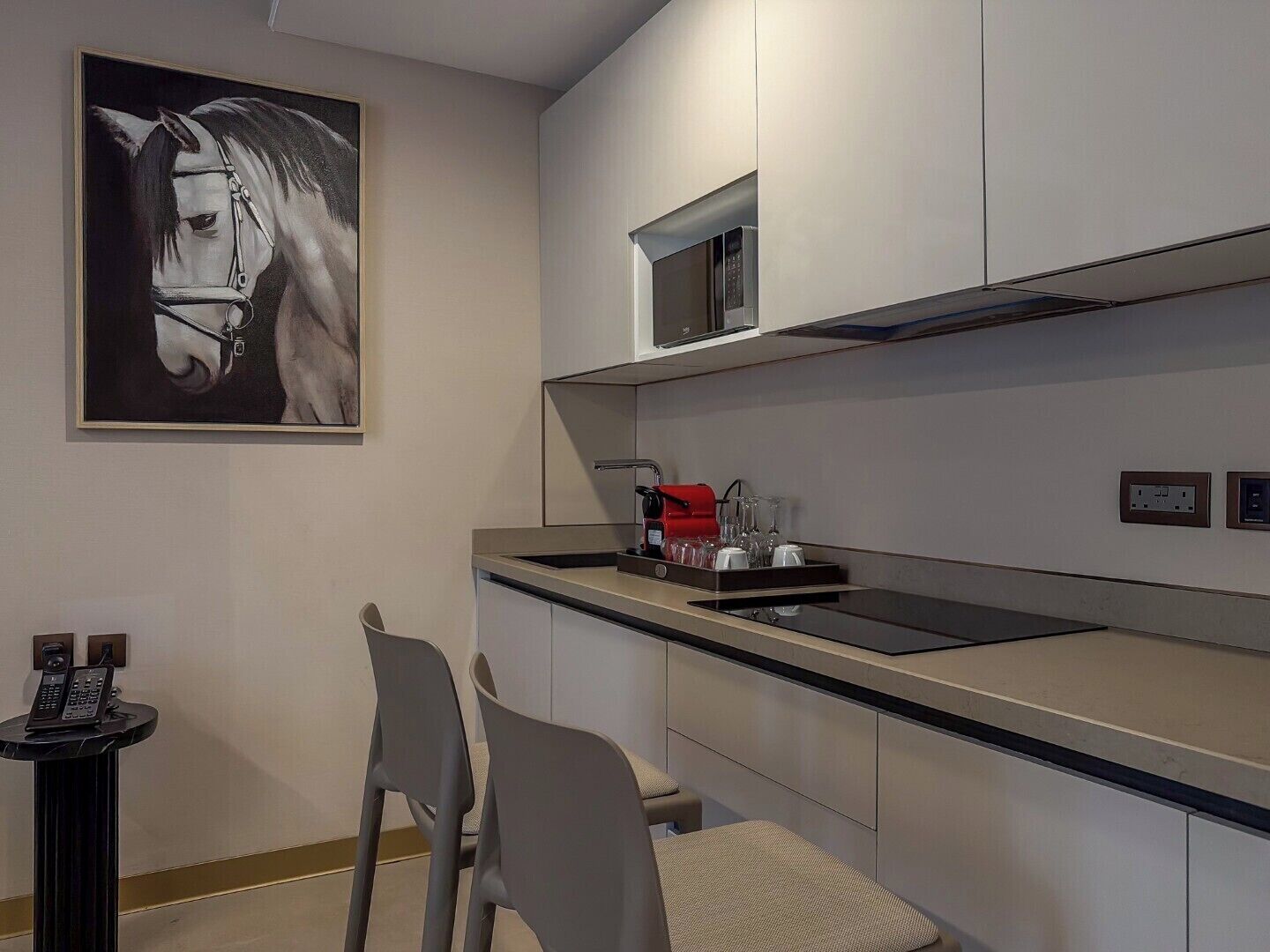 A modern kitchen with a microwave, a coffee maker, and a small sink. There are two chairs and a telephone on a small table.