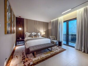 A modern bedroom with a large bed, a bench, a bedside table, a chair, and a view of the outdoors.