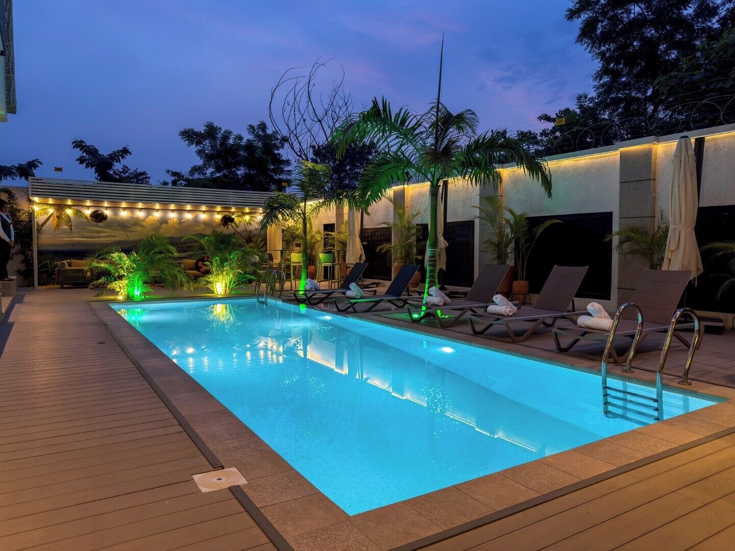 Una piscina con sillas de descanso y palmeras, rodeada por una terraza y una zona cubierta con luces colgantes.