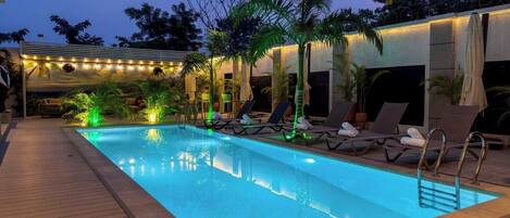Una piscina con sillas de descanso y palmeras, rodeada por una terraza y una zona cubierta con luces colgantes.