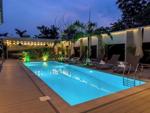 A swimming pool with lounge chairs and palm trees, surrounded by a deck and a covered area with string lights.