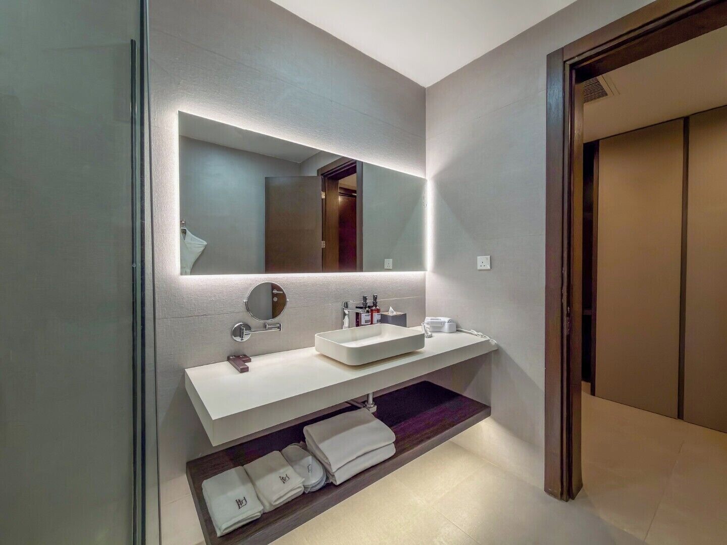 A modern bathroom with a large mirror, two sinks, a towel rack, and a shelf with folded towels.