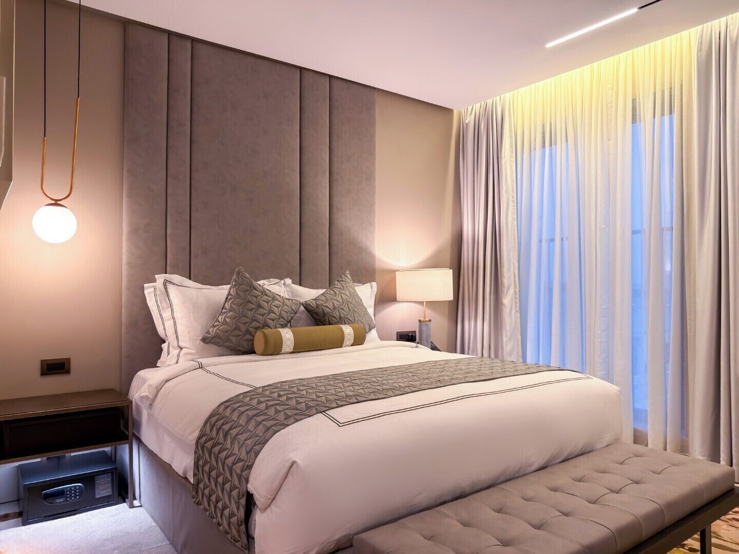 A modern hotel room with a large bed, bedside tables, a lamp, and a view of the outside through sheer curtains.