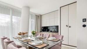 A modern dining room with a glass table, pink upholstered chairs, and a kitchen in the background. - Bluewave - Wave Resort & Spa (Międzyzdroje)