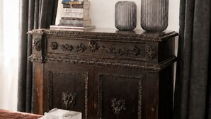 Ein Schlafzimmer mit einem braunen Bett, einer Holzkommode mit dekorativen Schnitzereien, einem Bücherstapel und einem gerahmten Bild an der Wand.