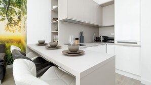 A modern kitchen with white cabinetry, a central island, and a dining area with a view of a tree through a window. - Nubo - Wave Resort & Spa (Międzyzdroje)
