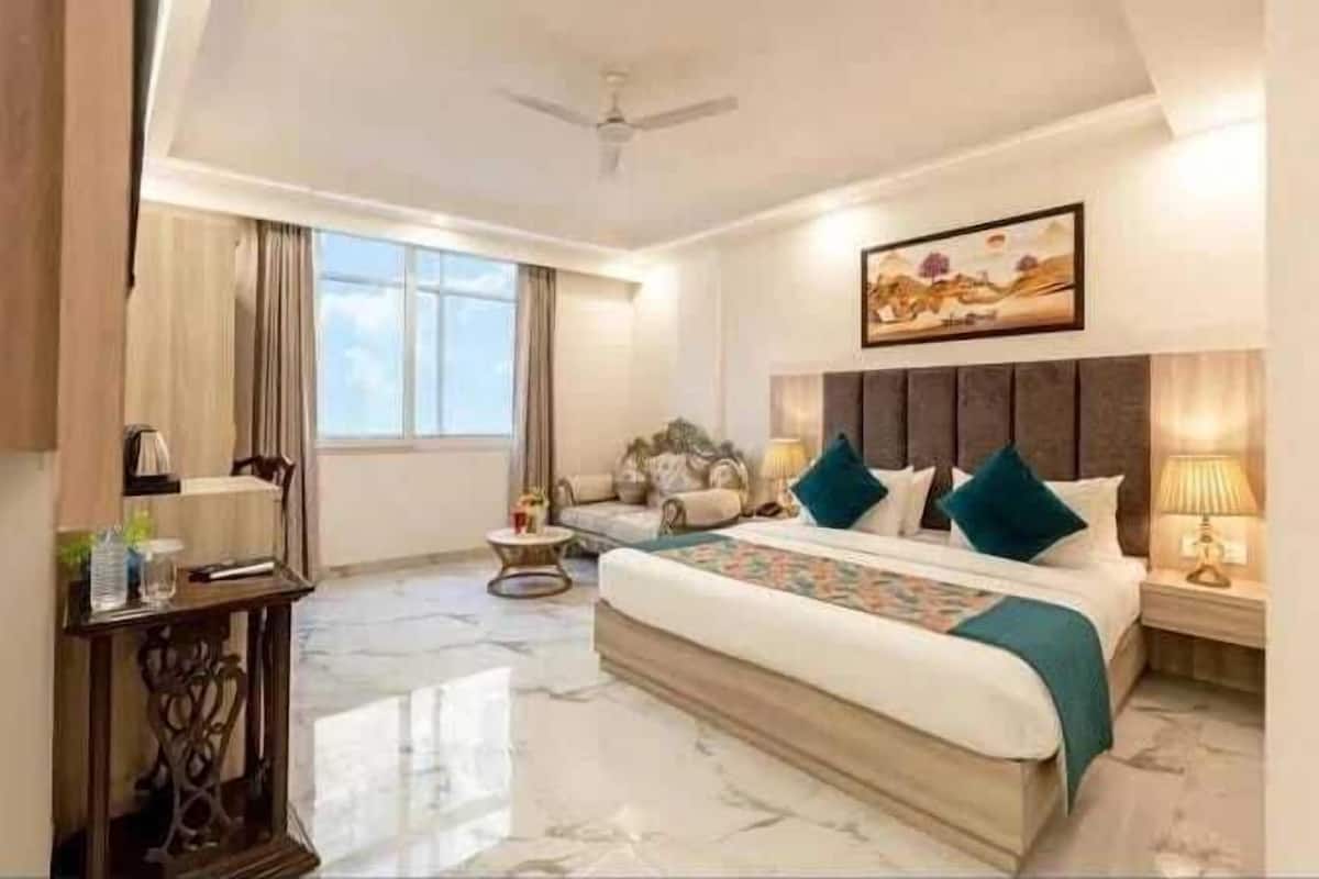 A hotel room with a large bed, a bedside table, a nightstand, a ceiling fan, a window with curtains, and a painting on the wall.