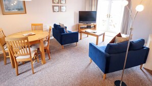 Ein Wohnzimmer mit Esstisch, blauen Sofas, Fernseher und Stehlampe.