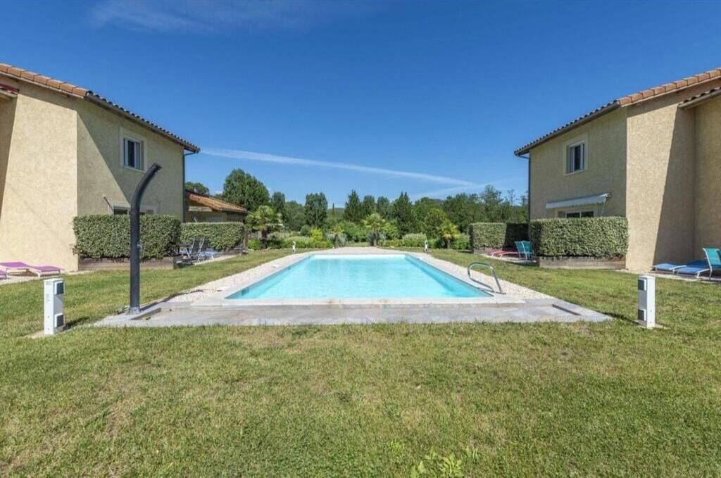 A swimming pool with a clear blue water, surrounded by a well-maintained lawn and two beige buildings with tiled roofs.