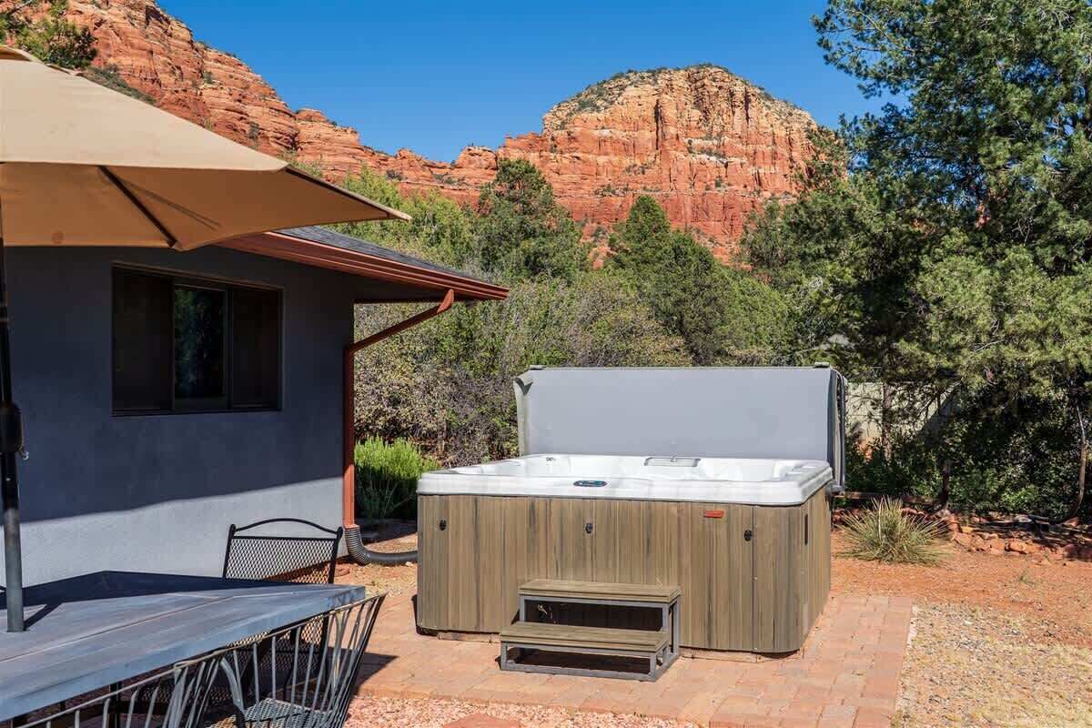 Un bain à remous extérieur avec un revêtement en bois, une table de patio et une chaise, le tout installé sur un fond de formations rocheuses rouges et de verdure.