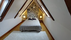 A bedroom with a sloped ceiling, stone wall, two bedside lamps, a bed with a grey duvet, and a white rug. - Charming 6-person house with large terrace - near Salers and Puy Mary (Anglards-de-Salers)