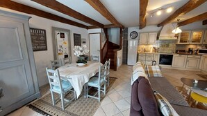 A dining area with a table and chairs, a sofa, a kitchen with cabinets and appliances, and a staircase. - Charming 6-person house with large terrace - near Salers and Puy Mary (Anglards-de-Salers)