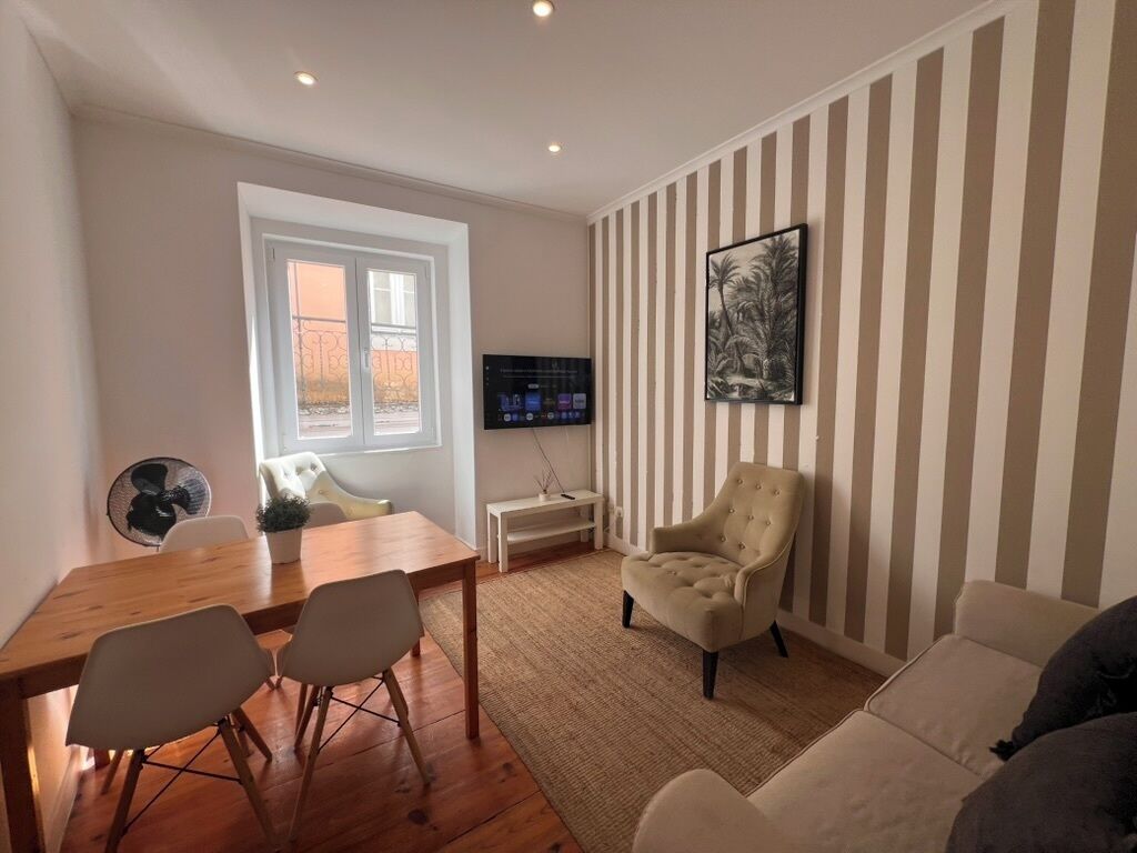 A living room with a wooden dining table, two chairs, a beige armchair, a sofa, a TV on a stand, a window with a view, and striped wallpaper.