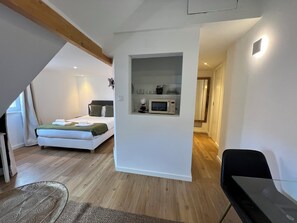 A bedroom with a bed, a small kitchenette, a chair, and a wooden ceiling beam.