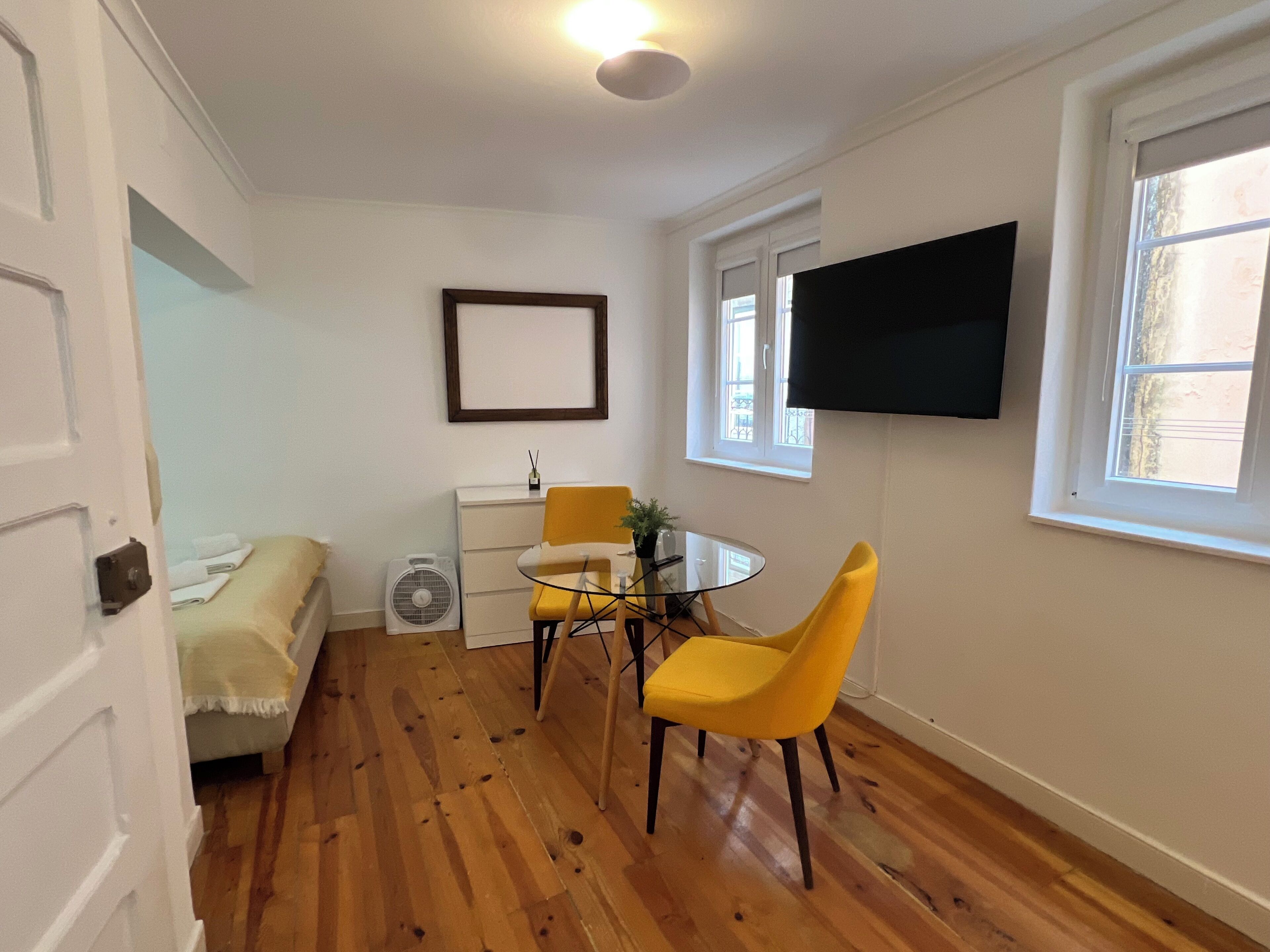 Una habitación con una cama, una mesita con sillas, un ventilador, un televisor en la pared y una ventana.