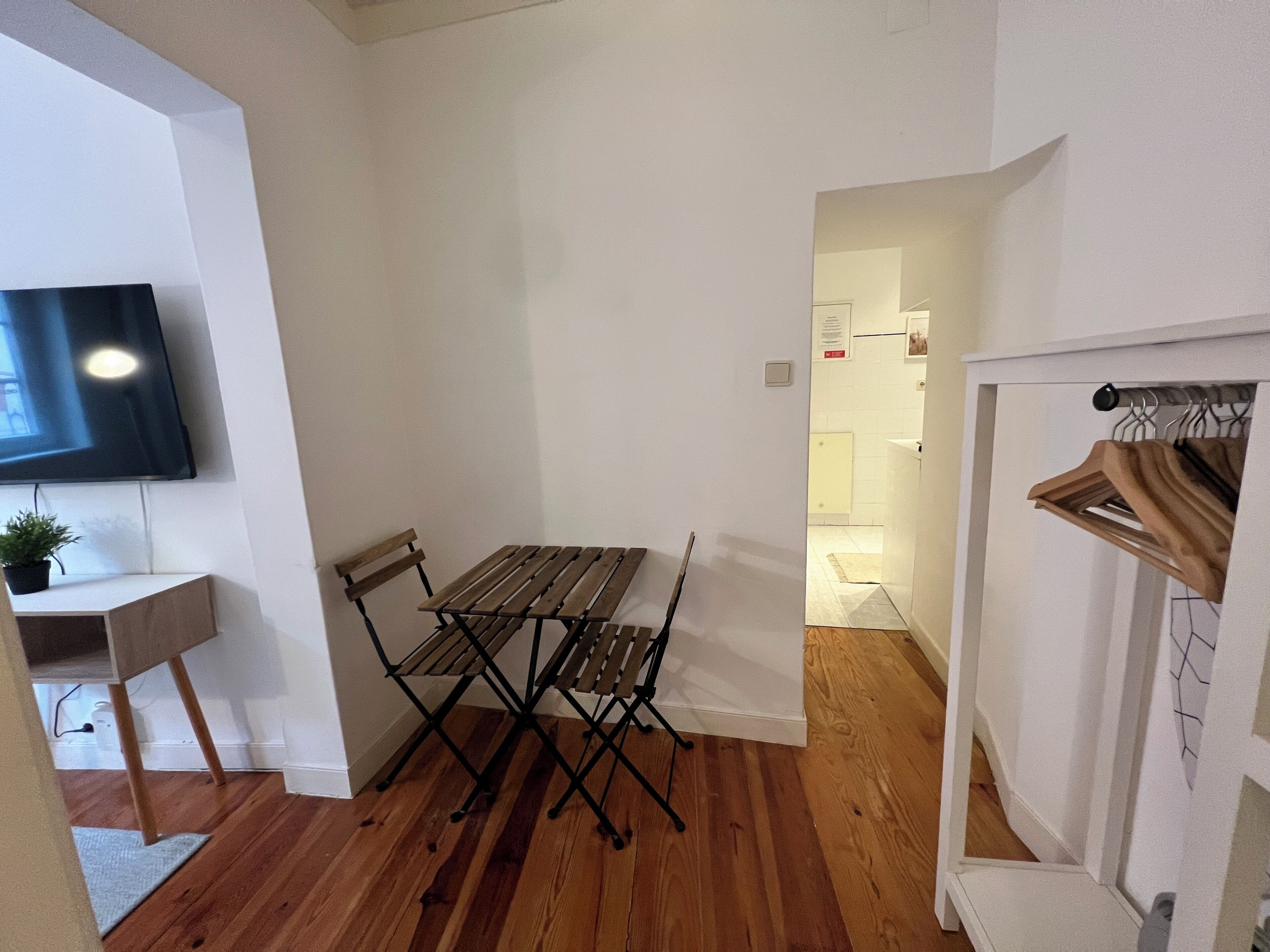 A compact living space with a dining table and chairs, a mounted TV, a small plant, and a coat rack.