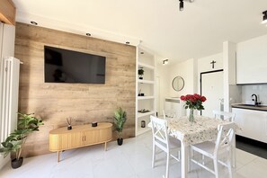 A modern kitchen with white cabinets, a wooden wall panel, a TV mounted above the counter, and a dining area with a table and chairs.