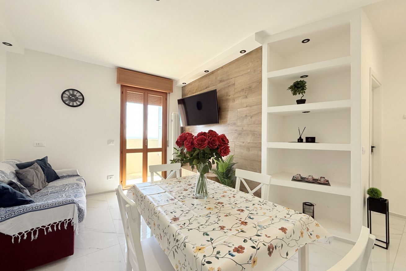 A modern living room with a sofa, a dining table with a floral tablecloth, a TV mounted on a wooden wall, and a bookshelf.