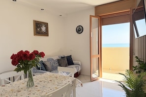 A room with a dining table, a sofa, a vase of red roses, a clock, and a view of the sea through an open door.