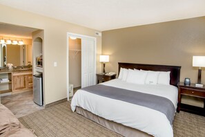 Habitación de hotel con cama, lámparas de noche, mesita de noche, escritorio con lámpara, espejo, armario y baño con lavabo y espejo.