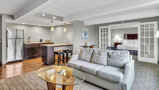 A modern living room with a glass coffee table, grey sofa, and a kitchen area with a refrigerator and bar stools. - Red Lion Inn & Suites Urbana (Urbana)