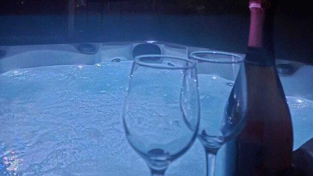 Un bain à remous avec deux verres et une bouteille, entouré d’arbres et d’un bâtiment.
